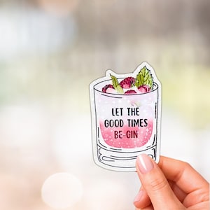 May include: A die-cut sticker featuring a cocktail glass filled with a pink liquid, raspberries, and mint leaves. The text "LET THE GOOD TIMES BE-GIN" is printed in black on the glass, which has a sparkling, glittery effect throughout the design.