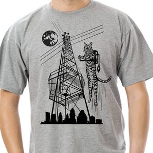 May include: A gray t-shirt with a black and white illustration of a cat hanging from a radio tower. The cat is wearing a backpack and holding a phone. The illustration also includes a city skyline and a crescent moon.