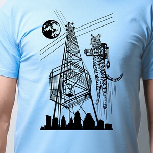 May include: A light blue t-shirt with a black and white illustration of a cat wearing a jetpack and flying past a radio tower. The cat is flying over a city skyline with a moon in the background.
