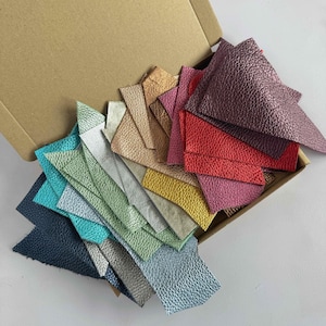 May include: A selection of leather swatches in various colours and textures, presented in a cardboard box. The swatches feature shades of blue, green, brown, red, and purple.