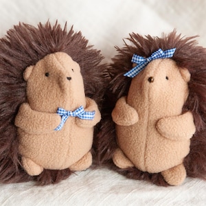 Könnte beinhalten: Zwei Stofftier-Igel. Sie haben beige Körper, braune Fellstacheln und kleine schwarze Augen. Einer hat eine blau-weiß karierte Schleife um den Hals, der andere eine Schleife auf dem Kopf. Auf weißem Hintergrund.
