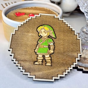 May include: Wooden coaster with a pixelated image of a character in green and yellow clothing. The coaster has a brown background and a white, jagged border. A set of coasters is visible in a round box.