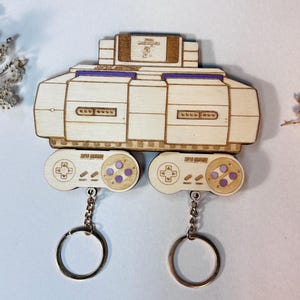 May include: Wooden keychain set featuring a Super Nintendo console and two controllers. The console is light brown with purple accents. The controllers have purple buttons and the text "SUPER NINTENDO". Each controller has a silver keychain ring.