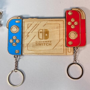 Puede incluir: Llavero de madera de Nintendo Switch con diseños de mando azul y rojo. La sección central muestra el logotipo y el texto de Nintendo Switch. Cada mando tiene un llavero plateado.