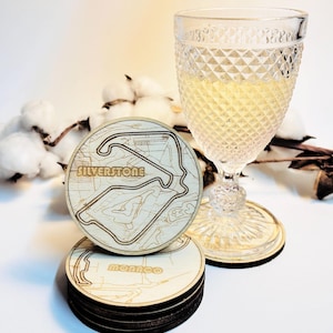 Könnte beinhalten: Holzuntersetzer mit hellem Holzfinish, einer mit dem Silverstone-Rennstreckendesign. Das Wort "Silverstone" ist auf dem Untersetzer eingraviert. Ein klares Glas mit Rautenmuster und einem Getränk steht daneben.
