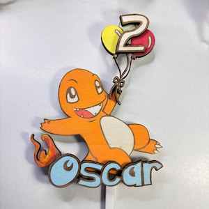 Puede incluir: Un adorno de pastel de madera para cumpleaños, con un personaje de dibujos animados naranja sosteniendo globos con el número 2. El personaje tiene una cola de llama y el nombre "Oscar" en letras azules. Los globos son amarillos y rojos.