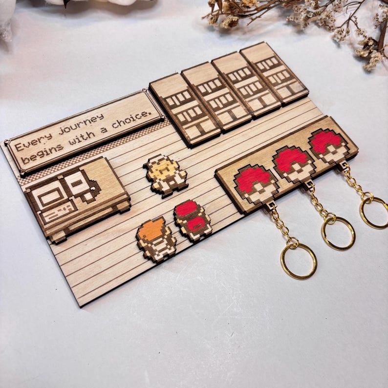 May include: Wooden display with pixelated game-themed designs. It features a game console, character figures, and red and white ball keyrings with gold chains. The text "Every journey begins with a choice" is displayed on the top left.