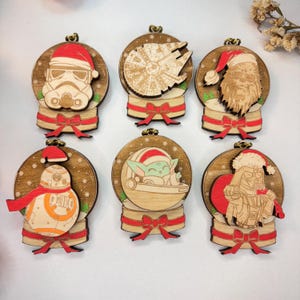 Op de afbeelding: Set van zes houten kerstversieringen met Star Wars-personages in sneeuwbolontwerpen. Elk ornament heeft een rode strik en een gouden lus om op te hangen. Personages zijn Stormtrooper, Darth Vader en Baby Yoda.