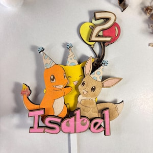 Puede incluir: Un adorno de pastel de madera para un cumpleaños, con tres personajes de dibujos animados con gorros de fiesta. Los personajes se abrazan, con el nombre "Isabel" debajo en letras rosas. Un número "2" está encima, sujeto a globos.