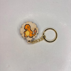 May include: Wooden keyring featuring a pixelated Charmander design. The Charmander is orange with a yellow belly and a red flame on its tail. The keyring has a gold-coloured chain and ring.