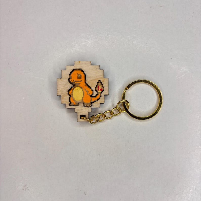 May include: Wooden keyring featuring a pixelated Charmander design. The Charmander is orange with a yellow belly and a red flame on its tail. The keyring has a gold-coloured chain and ring.