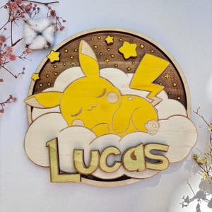 Può includere: Un decoro da parete in legno con un Pikachu giallo dei cartoni animati che dorme su una nuvola con uno sfondo di notte stellata marrone. Il nome "Lucas" è scritto in lettere gialle sotto la nuvola.