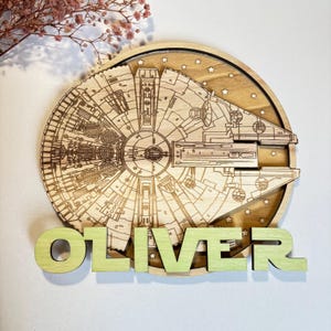 May include: A wooden wall decor featuring a laser-cut design of the Millennium Falcon spaceship from Star Wars. The spaceship is surrounded by a background of stars. The name "OLIVER" is written in green letters below the spaceship.
