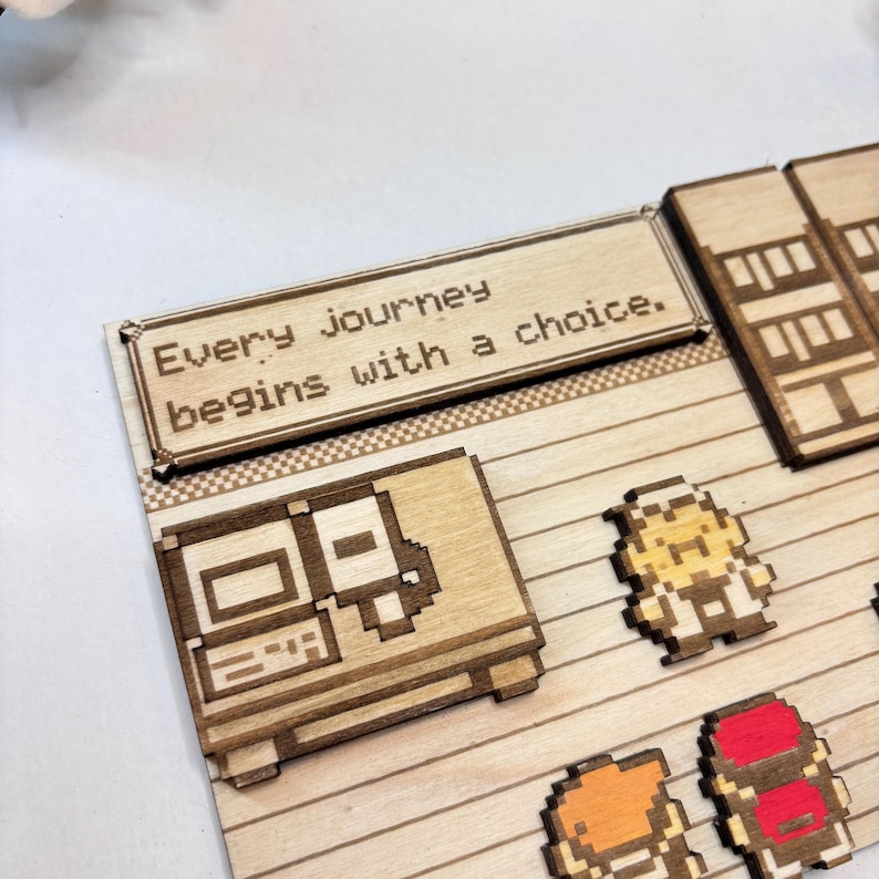May include: Laser-cut wooden pixel art with a computer, characters, and the text "Every journey begins with a choice." The pieces are light brown with darker etched details, set on a light wood background.