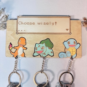 Peut inclure: Porte-clés en bois avec des motifs de Charmander, Bulbasaur et Squirtle. Le porte-clés est de forme rectangulaire avec le texte "Choose wisely!" au-dessus des personnages. Chaque personnage a un porte-clés attaché.