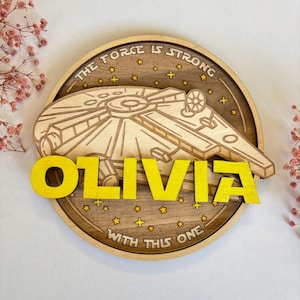 Puede incluir: Un letrero redondo de madera con una nave espacial amarilla inspirada en Star Wars y el texto "The Force Is Strong With This One". El nombre "Olivia" está escrito en letras amarillas debajo de la nave espacial.