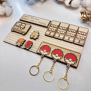 May include: A wooden laser-cut display with pixelated game-themed designs. It features a game console, character figures, and three red and white Pokeball keychains with gold-coloured chains and rings. The text "Every Journey begins with a choice" is also present.