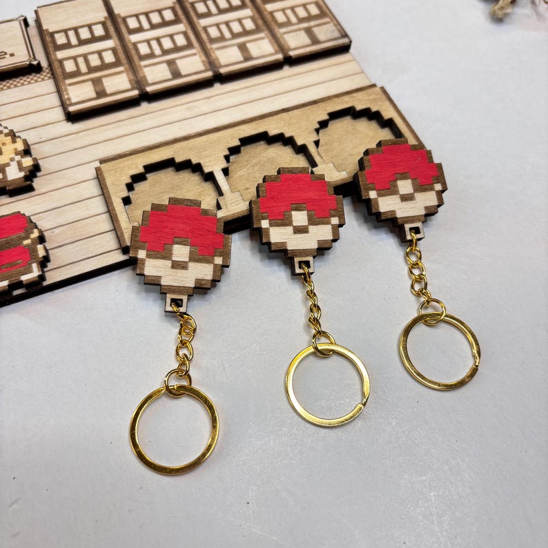 May include: Wooden keyrings with a pixelated design of a red and white ball. The keyrings have a gold-coloured chain and a round key ring. The keyrings are displayed on a wooden surface.