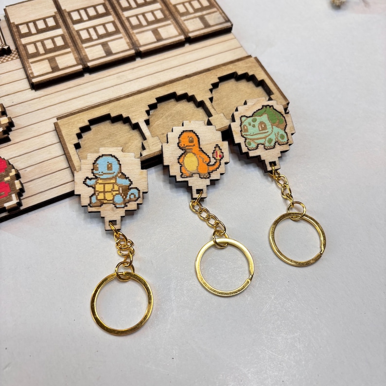 May include: Three wooden pixelated keychains featuring cartoon characters. The keychains have gold-coloured chains and rings. The characters are a blue turtle, an orange lizard, and a green plant-like creature. The keychains are on a wooden display.