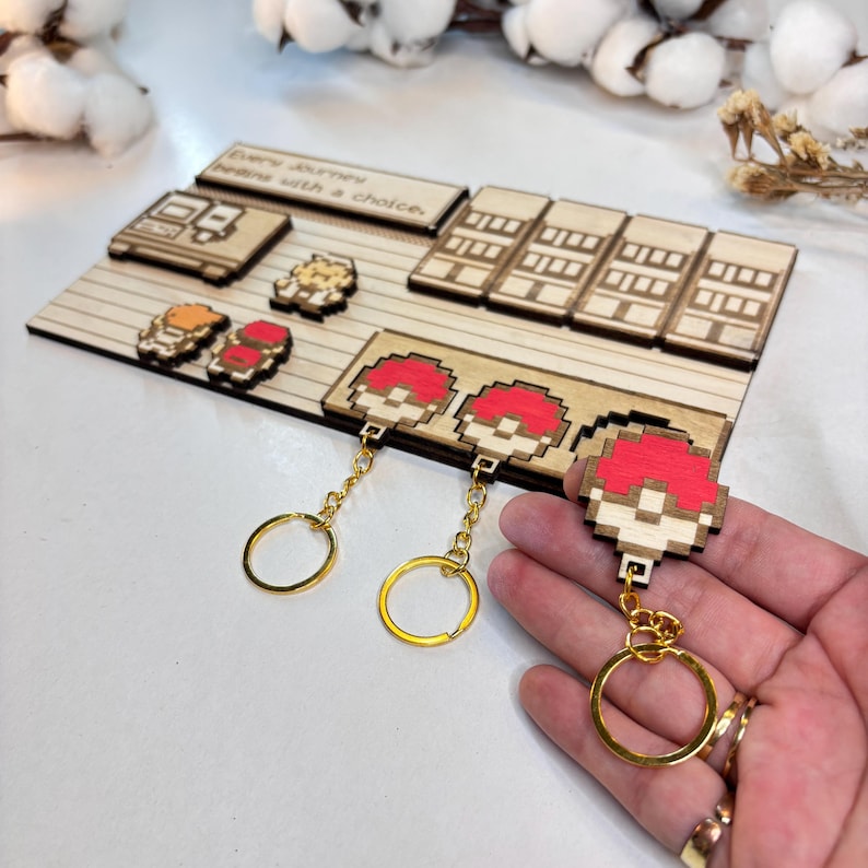 May include: A selection of wooden keychains featuring pixel art designs, including red and white mushroom shapes and other game-themed figures. The keychains have gold-coloured rings and chains. A wooden display board with the text "Every journey begins with a choice" is also visible.