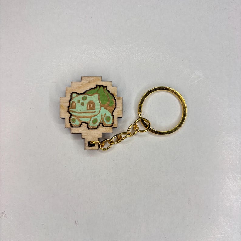May include: A wooden keychain with a pixelated Bulbasaur design. The Bulbasaur is coloured in shades of green and light green, with a dark green bulb on its back. The keychain has a gold-coloured chain and key ring.