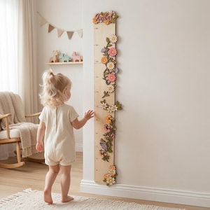Può includere: Un'asta di crescita in legno con decorazioni floreali e il nome "Isabel". Il grafico è decorato con fiori di legno colorati e misura in centimetri. Un bambino piccolo è vicino al grafico.