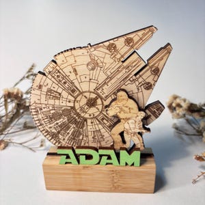 Peut inclure: Maquette en bois découpée au laser du Faucon Millenium, avec les figurines de Chewbacca et Han Solo. Le modèle est monté sur une base en bambou avec le nom "ADAM" en vert. Un objet sur le thème de Star Wars, avec des détails complexes.