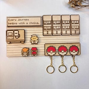 Op de afbeelding: Houten wanddecoratie met pixelontwerpen en de tekst "Every journey begins with a choice." Bevat een computer, personages en drie sleutelhangers met rood-witte balontwerpen. De sleutelhangers hebben goudkleurige ringen.
