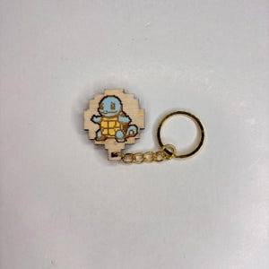 May include: Wooden keyring featuring a pixelated Squirtle design in blue and yellow. The keyring has a gold-coloured chain and ring. The Squirtle is set on a light-coloured wooden base.