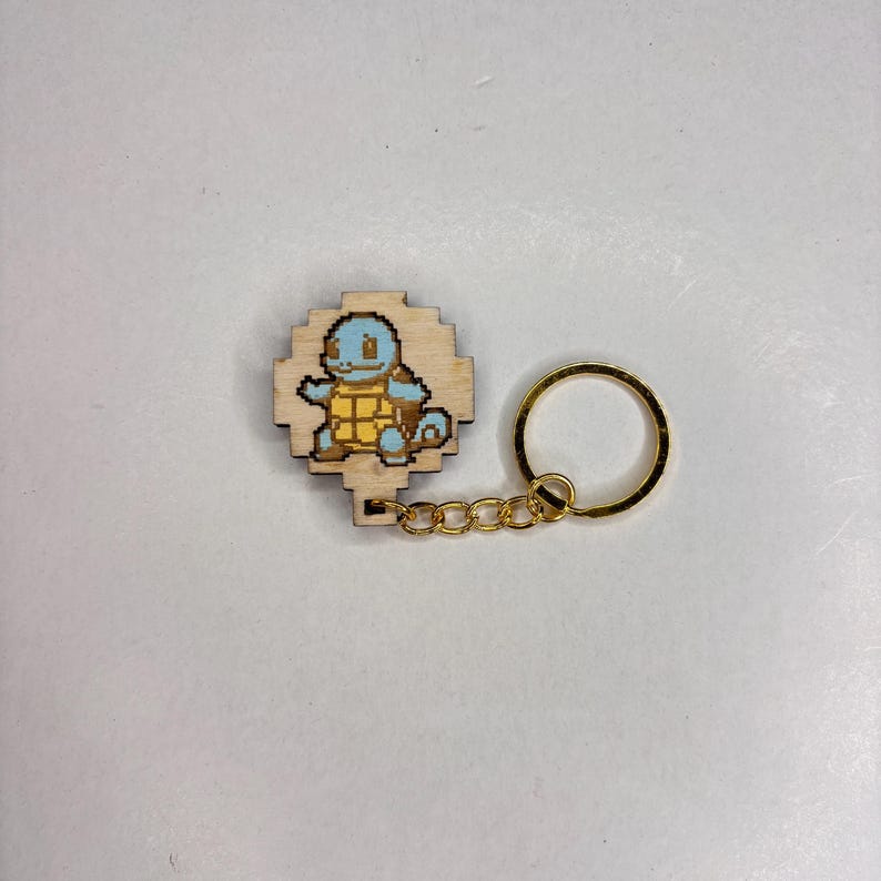 May include: Wooden keyring featuring a pixelated Squirtle design in blue and yellow. The keyring has a gold-coloured chain and ring. The Squirtle is set on a light-coloured wooden base.