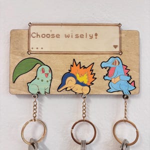 Könnte beinhalten: Schlüsselhalter aus Holz mit drei bunten Zeichentrickfiguren und Schlüsselanhängern. Der Text "Choose wisely!" steht über den Figuren. Der Schlüsselhalter ist aus hellbraunem Holz.