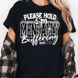 May include: Black t-shirt with the text "PLEASE HOLD I'M MENTALLY BUFFERING" in white. The text is in a bold, blocky font with a loading bar graphic. The shirt is a crew neck style.