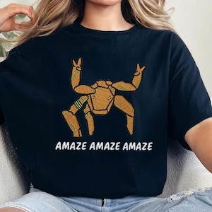 May include: A black short-sleeved t-shirt featuring a graphic of a golden-brown crab with raised claws. Below the crab, the text "AMAZE AMAZE AMAZE" is printed in white capital letters. The shirt has a crew neckline and a relaxed fit.