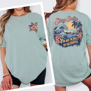 May include: A light green t-shirt with a floral design on the front and a graphic on the back that says "Stay Salty" with a wave, palm trees, and the text "You are the salt of the earth".
