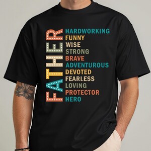 Peut inclure: T-shirt noir avec le mot « FATHER » en lettres colorées superposées sur le côté gauche. Le côté droit présente une liste d'attributs positifs : travailleur, drôle, sage, fort, courageux, aventureux, dévoué, intrépide, aimant, protecteur et héros.