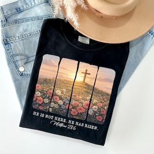 May include: Black t-shirt with a floral and cross design. The shirt features the text "HE IS NOT HERE. HE HAS RISEN. Matthew 28:6". The shirt is paired with light blue jeans and a beige hat.