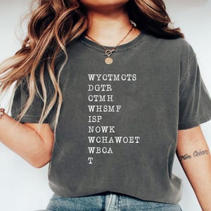 May include: Dark gray t-shirt with white text. The shirt features a list of capitalized letter combinations. The model is wearing a gold necklace and bracelet. The shirt is paired with blue jeans.