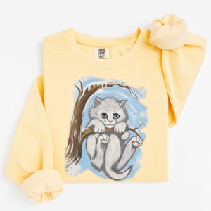 May include: A light yellow sweatshirt featuring a watercolor illustration of a gray kitten hanging from a tree branch. The kitten has blue eyes and pink paws. The background includes a blue sky and snowflakes.