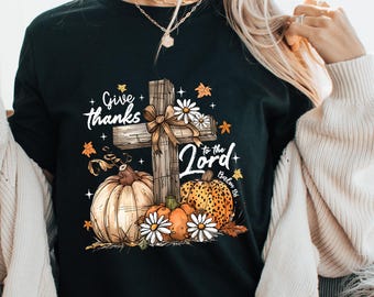 May include: Black t-shirt with a fall-themed graphic. The design features a wooden cross, pumpkins, daisies, and the text "Give thanks to the Lord". The shirt has a crew neck and short sleeves, perfect for autumn.