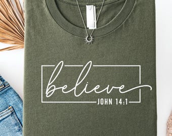 May include: An olive green t-shirt with the word "believe" in a white cursive font, inside a white rectangular frame. The text "JOHN 14:1" is below the word "believe". A silver necklace with a sunburst pendant is on the shirt.
