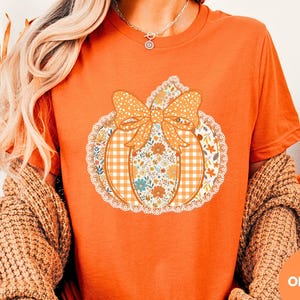 May include: Orange t-shirt featuring a pumpkin design with a bow, gingham, and floral patterns. The pumpkin is outlined with a white lace trim. The word "ORANGE" is in a circle at the bottom right. The shirt is part of a fall-themed collection.