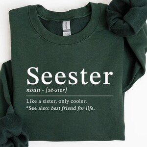 May include: A dark green sweatshirt with the word "Seester" in white, along with a definition. The definition reads "Like a sister, only cooler." The sweatshirt is folded on a white surface.