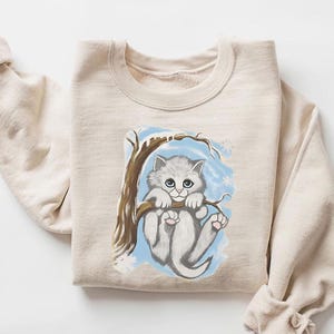 May include: Beige sweatshirt featuring a graphic of a gray kitten hanging from a tree branch. The kitten has blue eyes and pink paws, set against a blue and white snowy background. The sweatshirt has long sleeves with rolled cuffs.