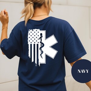 May include: Navy blue t-shirt with a white American flag and medical symbol graphic. The design includes a white star of life symbol with a caduceus. The shirt has a relaxed fit.