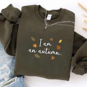 May include: An olive green sweatshirt with the text "I am an autumn" in white, decorated with autumn leaves. A silver chain necklace is draped across the sweatshirt. The sweatshirt is folded on top of a pair of blue jeans.