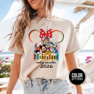 May include: Beige t-shirt featuring a Disneyland graphic with Mickey Mouse, Minnie Mouse, Donald Duck, and Goofy. The design includes a castle, fireworks, and the text "Disneyland Family Vacation 2026".
