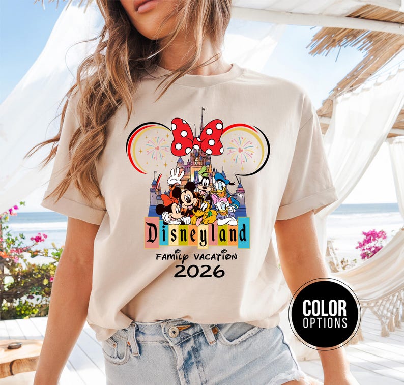 May include: Beige t-shirt featuring a Disneyland graphic with Mickey Mouse, Minnie Mouse, Donald Duck, and Goofy. The design includes a castle, fireworks, and the text "Disneyland Family Vacation 2026".