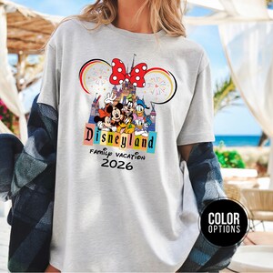 May include: A light gray t-shirt featuring a Disneyland design with Mickey Mouse, Minnie Mouse, Donald Duck, and other characters. The design includes a castle, fireworks, and the text "Disneyland Family Vacation 2026."