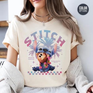 Może przedstawiać: Kremowa koszulka z grafiką Stitcha z Lilo & Stitch. Projekt zawiera Stitcha trzymającego brązowy przedmiot, ze słowem "STITCH" w czcionce gradientowej. Dodatkowy tekst brzmi "Session Surfing" i "Santa Monica Beach".