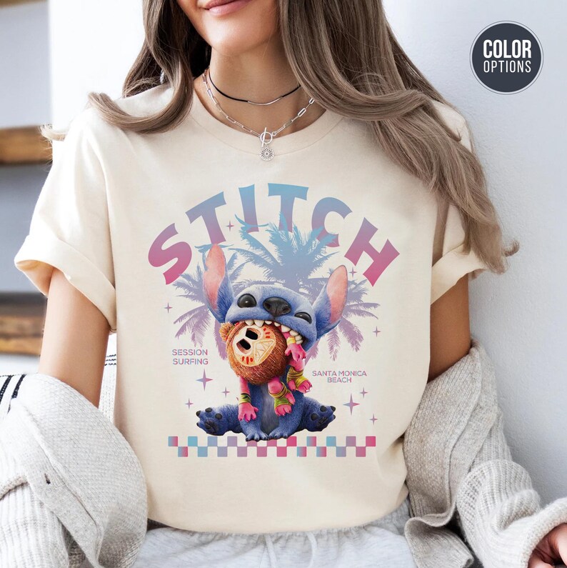 Może przedstawiać: Kremowa koszulka z grafiką Stitcha z Lilo & Stitch. Projekt zawiera Stitcha trzymającego brązowy przedmiot, ze słowem "STITCH" w czcionce gradientowej. Dodatkowy tekst brzmi "Session Surfing" i "Santa Monica Beach".